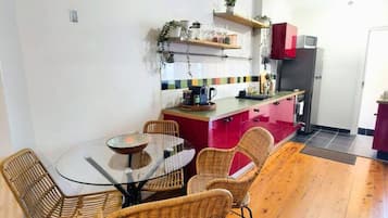 Zona pranzo con tavolo rotondo in vetro e sedie in vimini, cucina con frigorifero e pensili rossi, e pavimento in legno.