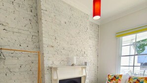 Ein Schlafzimmer mit weißer Ziegelwand, Kamin, einem Bett mit bunten Kissen, einem kleinen Tisch und einer Pendelleuchte.