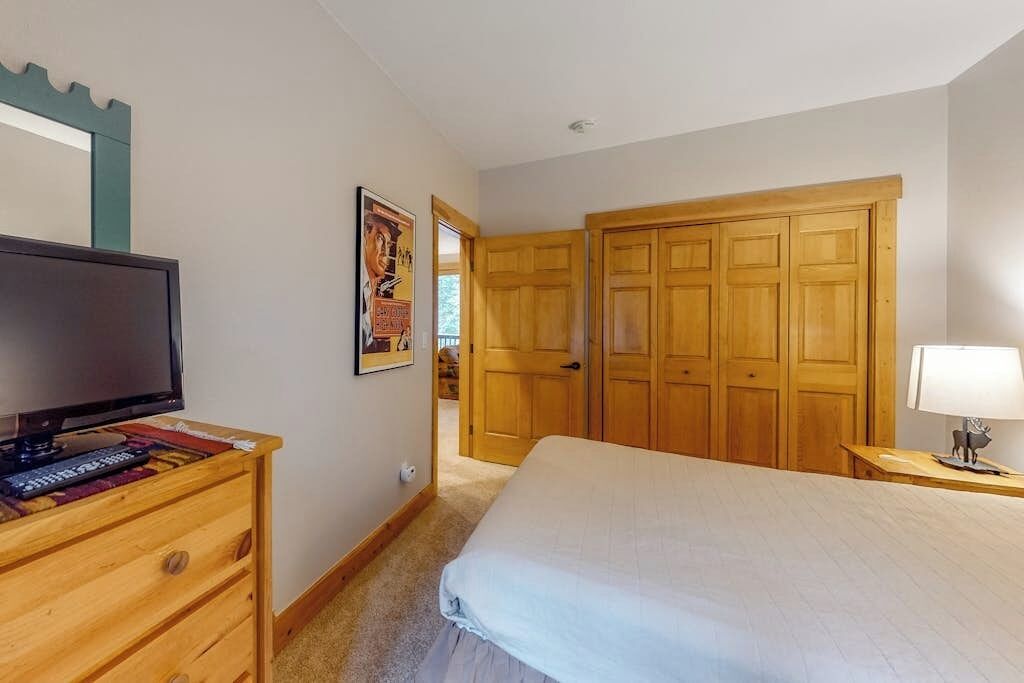 A bedroom with a bed, a dresser, a television, a wooden wardrobe, and a door leading to another room.
