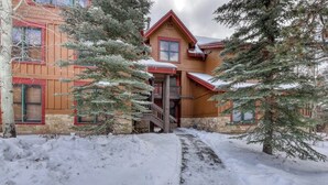 A wooden lodge with a central staircase, surrounded by snow-covered ground and trees.