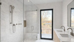 Une salle de bain moderne dotée d’une douche à parois de verre, d’une grande fenêtre et d’un meuble-lavabo en bois avec deux vasques.