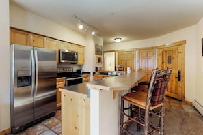 A kitchen with wooden cabinets, stainless steel appliances, a bar area, and a view of the living area. - Corner condo with hot tub & private washer/dryer - steps from the slopes (Keystone)