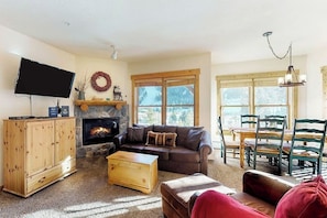 A living room with a fireplace, a television, a dining area with a table and chairs, and a large window. - Corner condo with hot tub & private washer/dryer - steps from the slopes (Keystone)