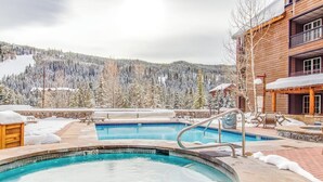 A pool area with two hot tubs, wooden deck, and a view of snow-covered trees and mountains. - Spacious, resort condo w/private balcony, shared pool & hot tub. Walk to slopes (Keystone)