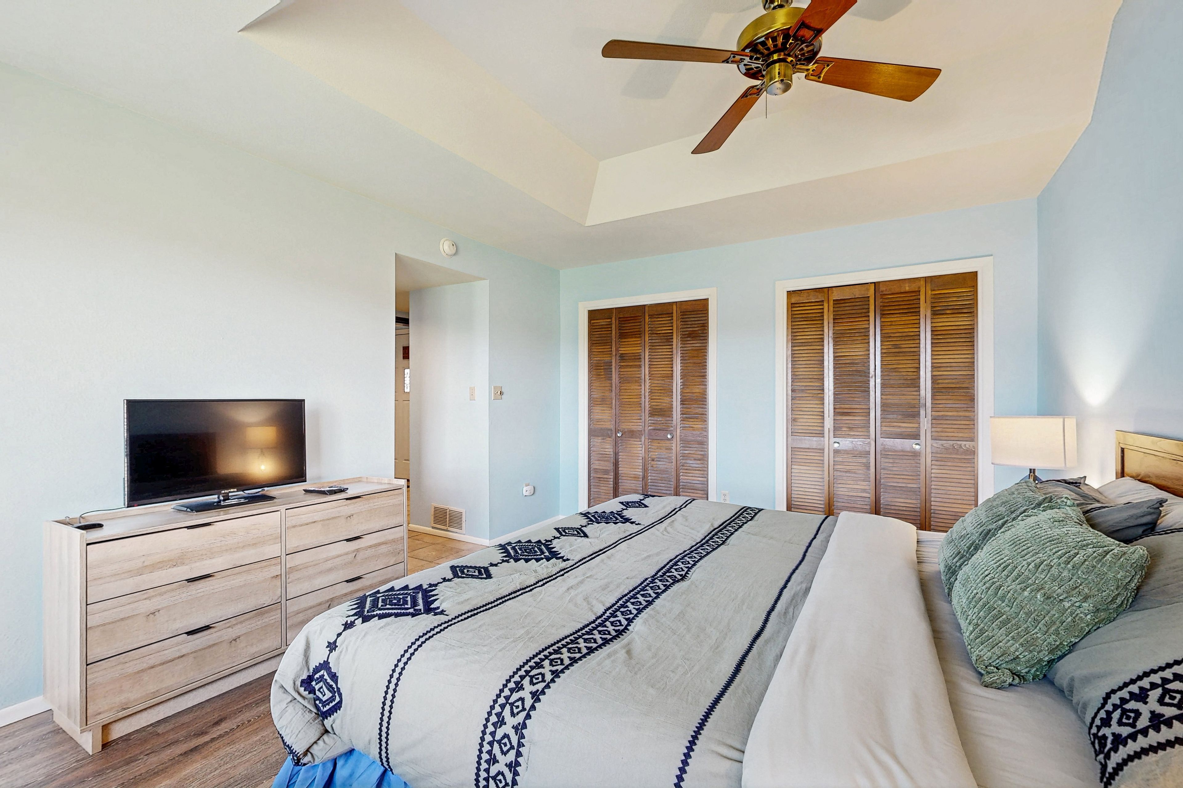 A bedroom with a bed, a television on a wooden dresser, a ceiling fan, and wooden shutters on the windows.