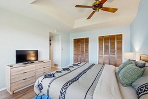 Un dormitorio con cama, televisor sobre un aparador de madera, ventilador de techo y persianas de madera en las ventanas.