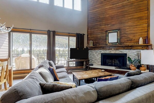 Ein Wohnzimmer mit einer großen Couch, einem Kamin, einem Fernseher und Blick ins Freie.