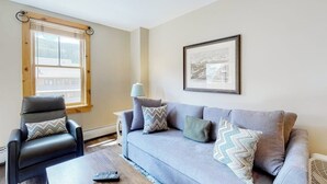 En stue med en grå sofa, en sort læderlænestol, et lille bord, en lampe og et indrammet billede på væggen.