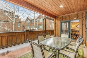 Un espace de restauration extérieur abrité, avec une table en verre et des chaises en métal, une terrasse en bois et une vue sur des immeubles résidentiels.