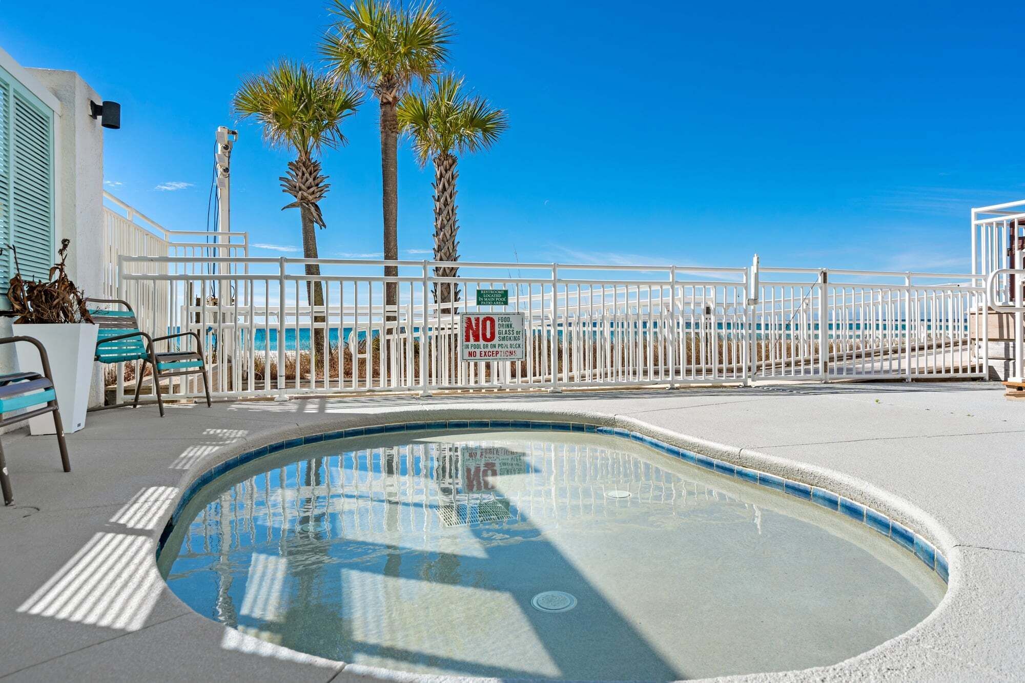 Une piscine entourée d’une clôture blanche, de palmiers et surplombée d’un ciel bleu azur.
