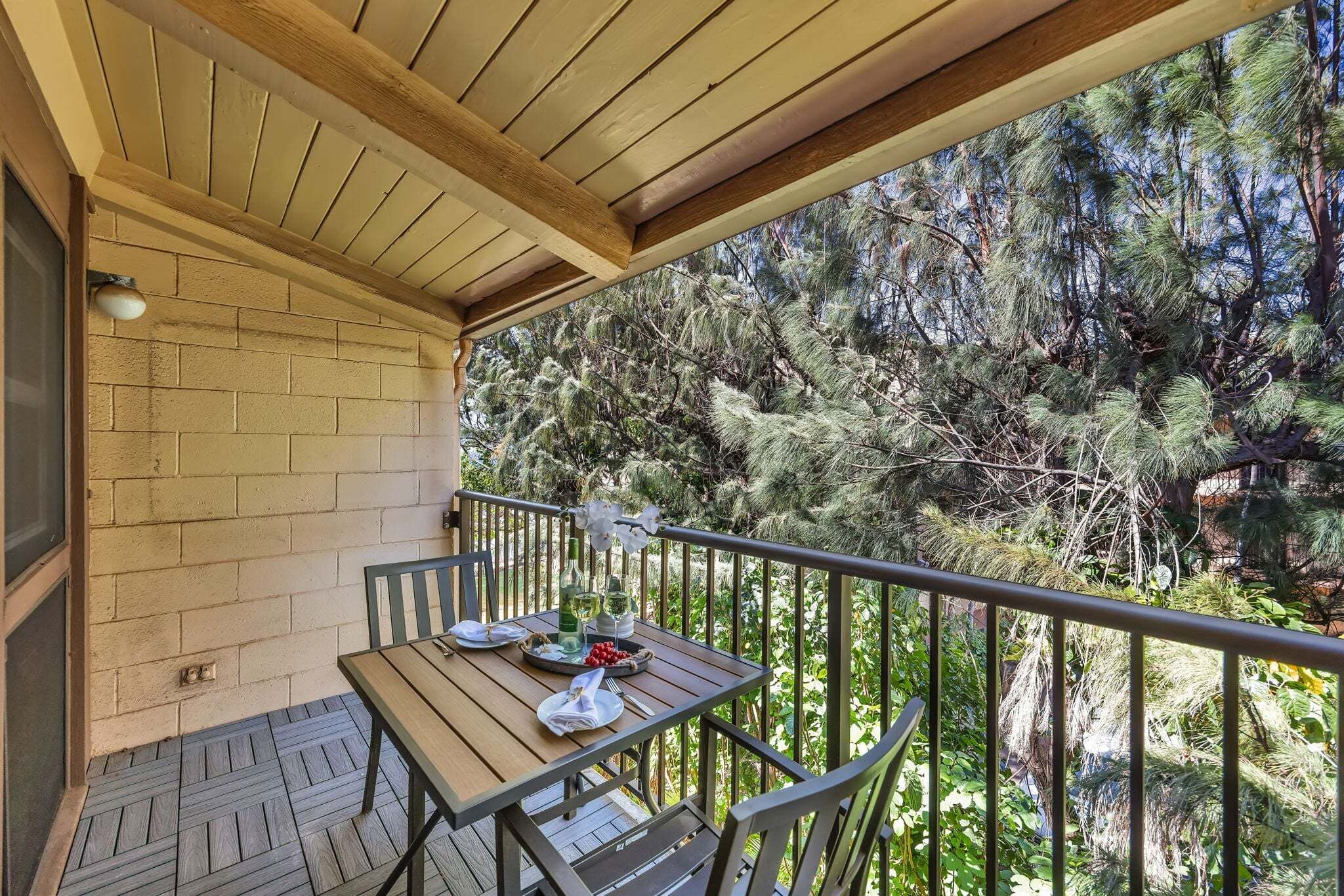 A wooden balcony with a table set for two, surrounded by metal railings, overlooking a dense forest.