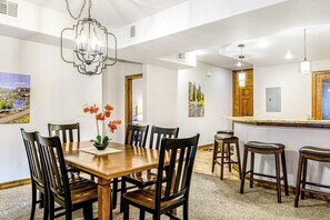 Zona pranzo con tavolo in legno, sedie, un lampadario e un bancone cucina con sgabelli.