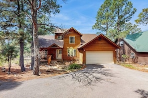 A two-story log cabin with a garage, surrounded by pine trees.