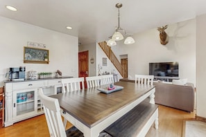 A dining area with a wooden table, white chairs, a coffee station, a television, and a mounted deer head.