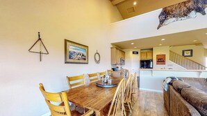 Un coin repas avec une table et des chaises en bois, une cuisine à l’arrière-plan et un escalier menant à un niveau supérieur.