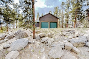 A cabin with two green garage doors, surrounded by a rocky terrain and tall pine trees. - Log Home with Mountain Views, Firepit & Gas Fireplace - Quick Drive to Skiing (Tabernash)
