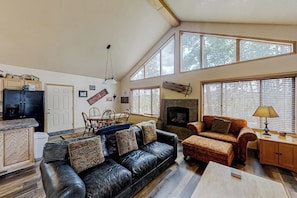 A spacious living room with a fireplace, a black leather sofa, a wooden dining table with chairs, and large windows. - Log Home with Mountain Views, Firepit & Gas Fireplace - Quick Drive to Skiing (Tabernash)