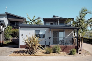 A single-story house with a covered porch, a small garden with various plants, and a clear blue sky. - Brooman 9 - Self-Contained Cabin with Covered Deck (Mollymook)