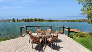 Un terrazzo in legno con un tavolo rotondo e sedie, con vista su un lago e una riva distante.
