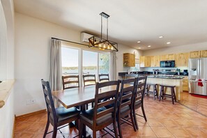 A dining area with a table and chairs, a kitchen with modern appliances, and a view through large windows. - Stunning lake house with dock, firepit, grill, canoes, & mountain views (Pagosa Springs)