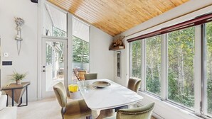 Un comedor con mesa y sillas, techo de madera y ventanas amplias con vista a árboles.