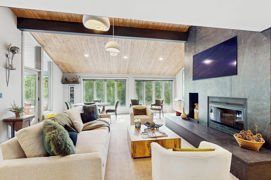 A spacious living room with a high ceiling, large windows, a fireplace, and a flat-screen TV mounted on the wall.