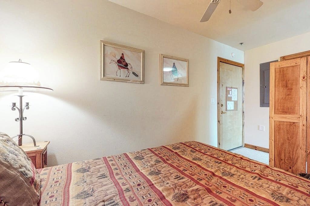 A bedroom with a bed, a lamp, two framed pictures, a wooden door, and a ceiling fan.