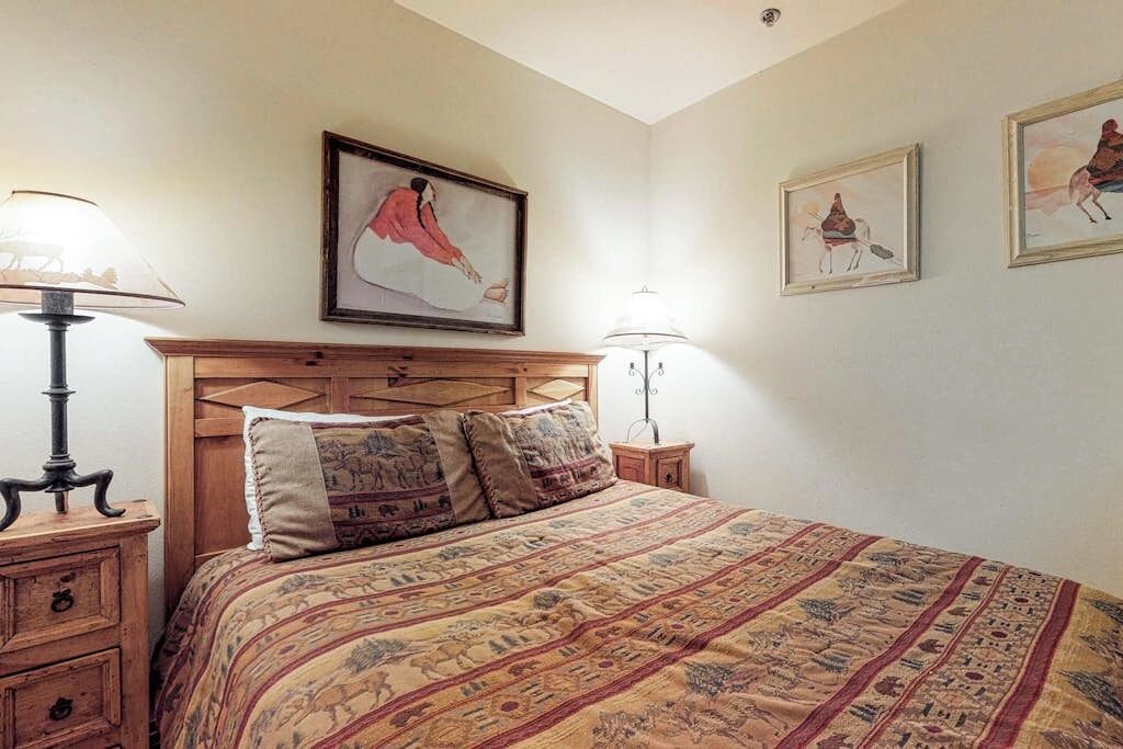 A bedroom with a wooden headboard, a patterned bedspread, two bedside lamps, and framed artwork on the wall.