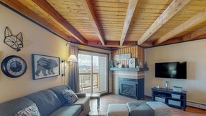 A living room with a fireplace, a sofa, ottomans, a TV, and a wall clock. - Conveniently Located Condo with Pool, Hot Tub & Sauna - Near Slopes & Lake (Silverthorne)