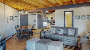 A spacious living area with a sofa, ottomans, a dining table with chairs, a kitchen in the background, and a wooden ceiling. - Conveniently Located Condo with Pool, Hot Tub & Sauna - Near Slopes & Lake (Silverthorne)