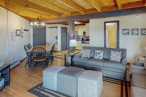 A spacious living area with a sofa, ottomans, a dining table with chairs, a kitchen in the background, and a wooden ceiling. - Conveniently Located Condo with Pool, Hot Tub & Sauna - Near Slopes & Lake (Silverthorne)
