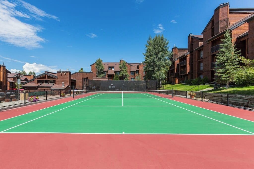 Una cancha de tenis con red, rodeada de edificios de ladrillo y árboles.