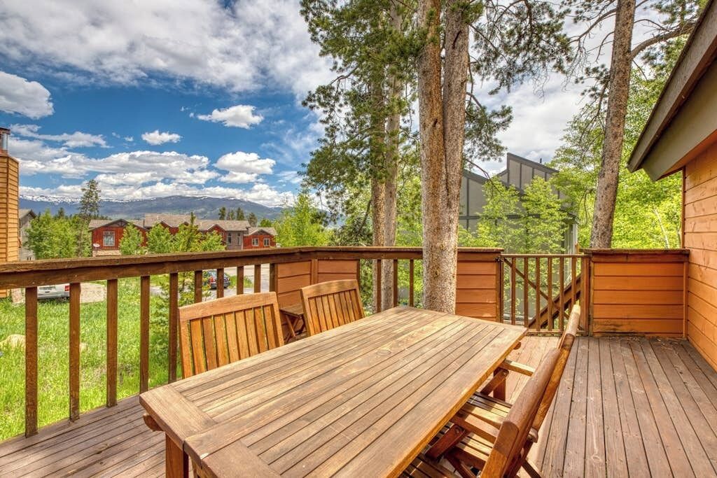 Eine Holzterrasse mit Tisch und Stühlen, umgeben von Bäumen und Blick auf Berge.