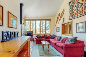 Ein Wohnzimmer mit einem roten Sofa, einem Holztisch und einem Fernseher. Es gibt große Fenster und einen Kamin.