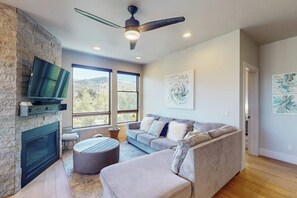 Ein modernes Wohnzimmer mit Kamin aus Stein, einem Flachbildfernseher, einem Deckenventilator und einem großen Fenster mit Blick ins Freie.