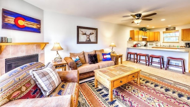 A living room with a sofa, coffee table, and patterned rug. There is a fireplace, a ceiling fan, and a kitchen area with stools. - Mountain family townhome w/ fireplace, private balcony & beautiful views (Silverthorne)