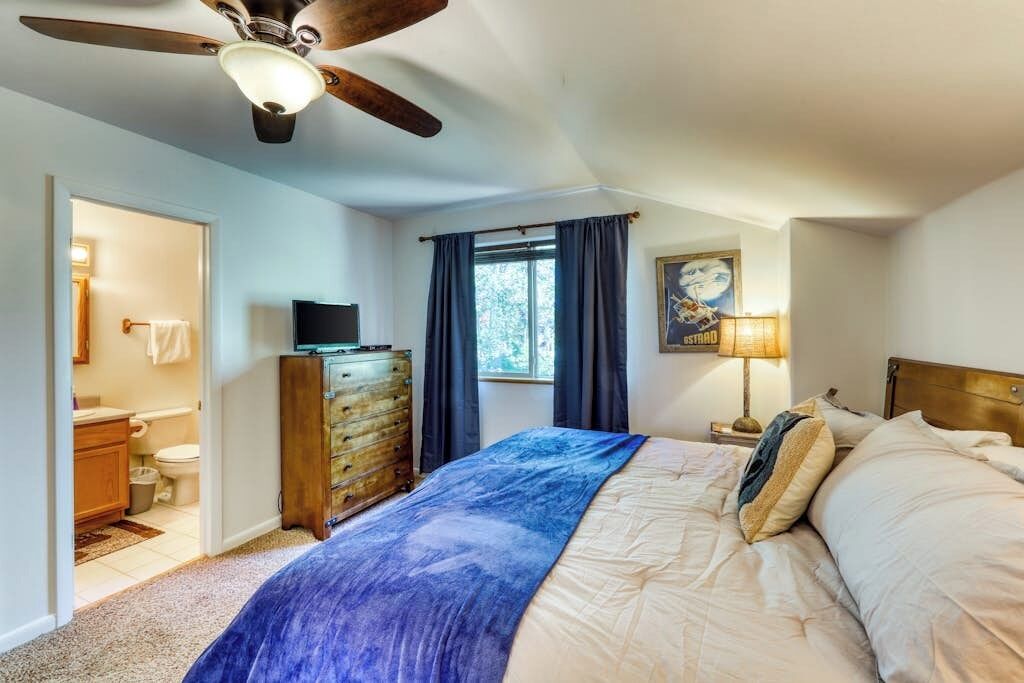 A bedroom with a bed, a ceiling fan, a dresser, a window with curtains, a painting, and a nightstand with a lamp.