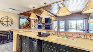 Une cuisine dotée d’un grand îlot central, de poutres en bois au plafond, de luminaires suspendus, d’une cheminée en pierre et d’une horloge murale.