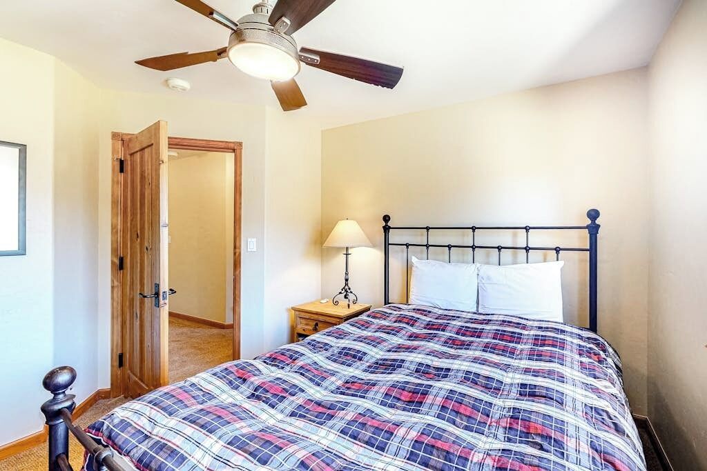 A bedroom with a bed, a ceiling fan, a nightstand with a lamp, and a door.