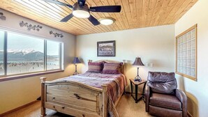 Une chambre avec un lit, une chaise, un ventilateur de plafond et une fenêtre donnant sur la montagne.