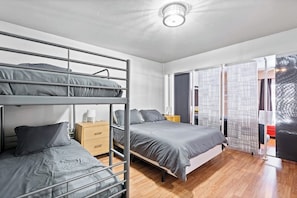 Ein Schlafsaalzimmer mit Stockbetten, einer Holzkommode, einem Nachttisch mit Lampe und einer Glastür zum Balkon.