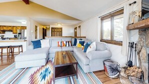Een woonkamer met een hoekbank, een houten salontafel, een kamerkleed met een patroon en een stenen open haard.