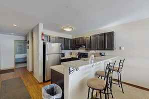 A compact kitchen with dark cabinets, a refrigerator, a stove, and a bar area with stools. - Lake & ski view condo | Pool, hot tub, sauna & cozy fireplace (Dillon)
