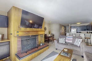 A living room with a fireplace, a sofa, a coffee table, and a kitchen area in the background. - Lake & ski view condo | Pool, hot tub, sauna & cozy fireplace (Dillon)