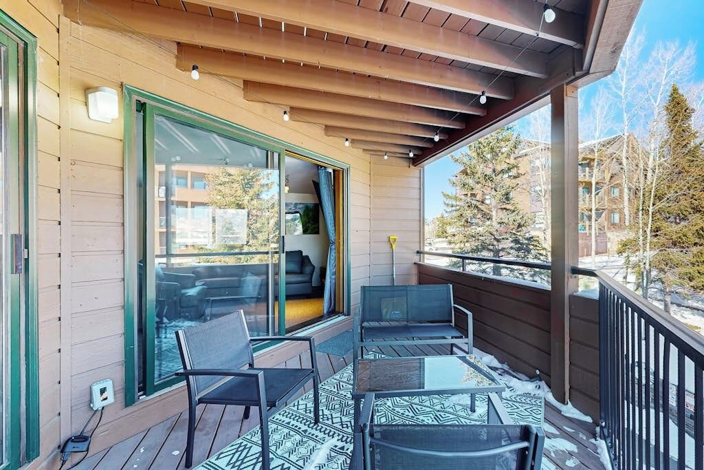 A wooden deck with outdoor furniture, a glass door leading to an interior space, and a view of a residential area with trees and buildings.