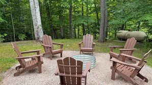 Eine runde Sitzgruppe mit Adirondack-Stühlen um ein Feuerkorb in einer bewaldeten Gegend.