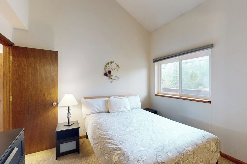 A bedroom with a bed, a nightstand, a lamp, a wooden door, and a window.