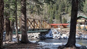 Un puente de madera cruza un río, rodeado de árboles y una cabaña con techo rojo.