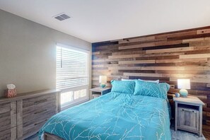 Ein Schlafzimmer mit hölzernem Kopfteil, einem Bett mit blauer Bettdecke, Nachttischen mit Lampen und einem Fenster mit Jalousien.