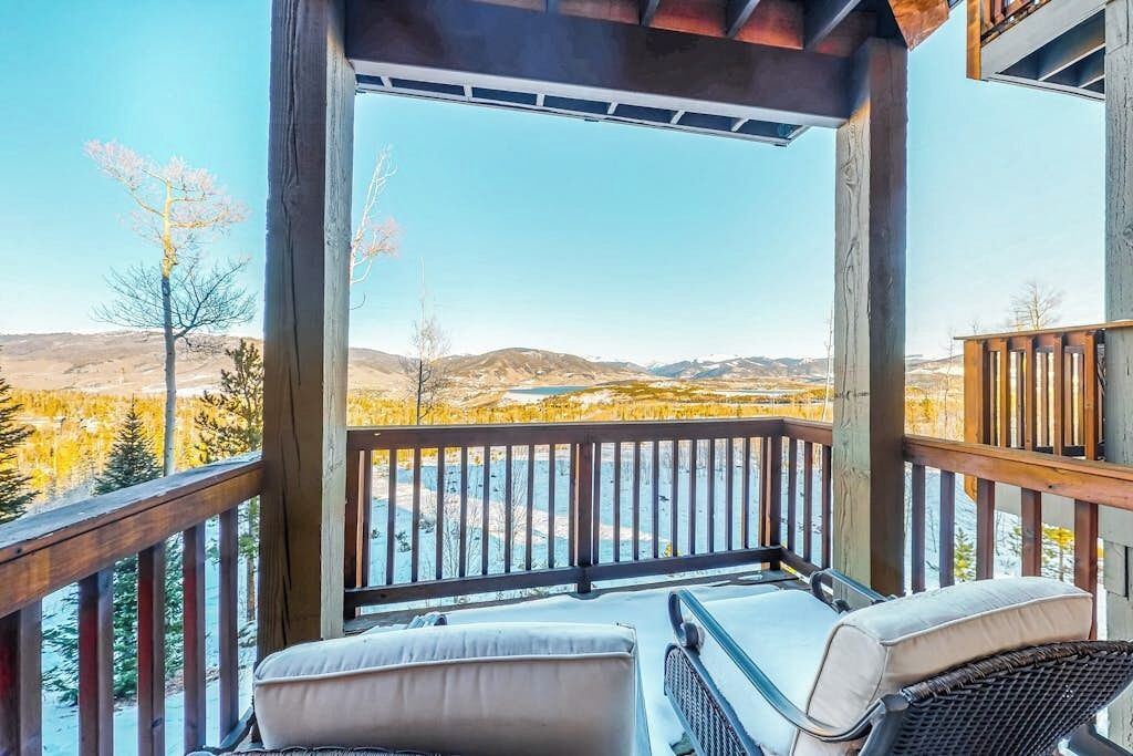 Una terraza de madera con dos sillas, una mesa y vistas a un valle con árboles y montañas.